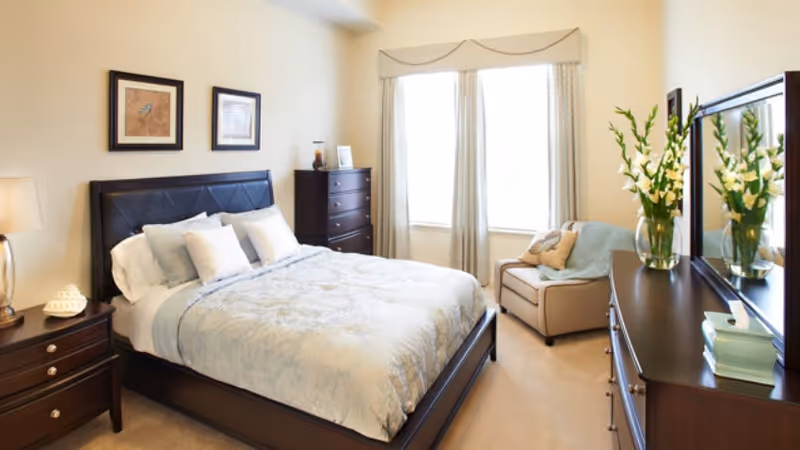 A well-lit bedroom with a large bed featuring a dark wooden frame and light-colored bedding. There are two framed pictures on the wall above the bed, a nightstand with a lamp and decorative items, a tall dresser, a comfortable armchair near a window with curtains, and a dresser with a mirror and a vase of flowers.