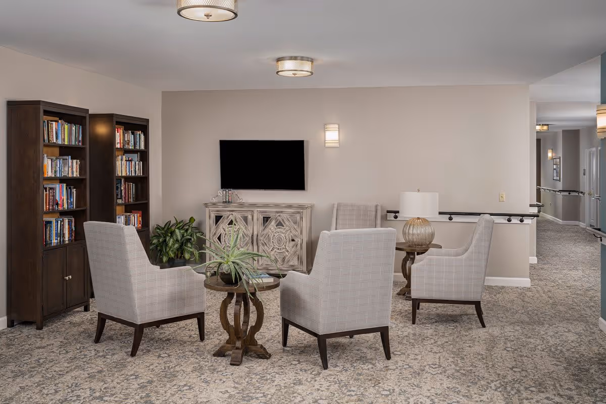 A cozy sitting area with four upholstered armchairs arranged around two small wooden tables with plants on them. Behind the chairs is a decorative cabinet with a flat-screen TV mounted on the wall above it. To the left, there are two tall bookshelves filled with books. The room has beige walls, patterned carpet, and ceiling lights. A hallway is visible to the right.
