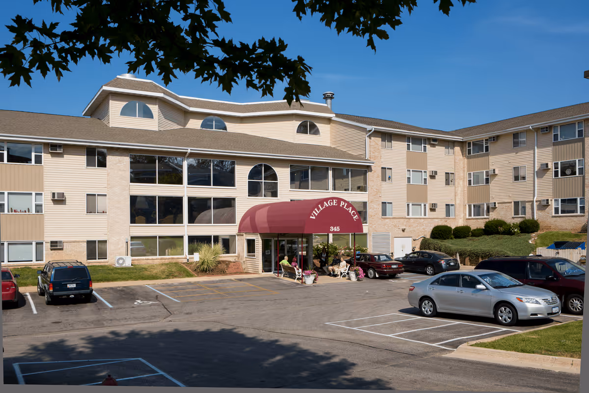 Exterior view of Village Place, a multi-story senior living facility with beige siding and large windows. A maroon awning with the name 'Village Place' and the number '345' covers the main entrance where several people are sitting on benches. Several cars are parked in the parking lot in front of the building, and tree branches frame the top of the image.