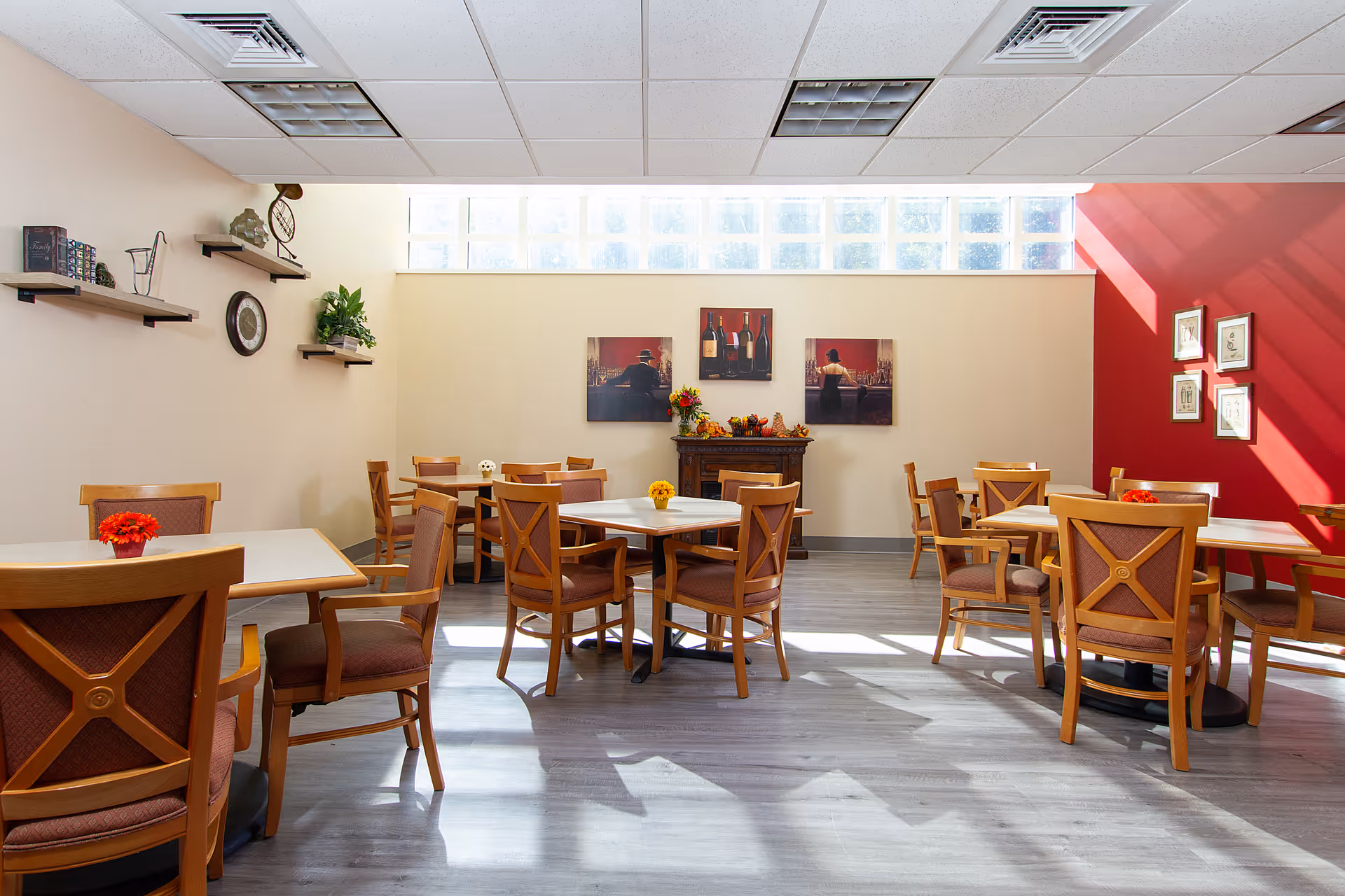 A bright dining room with several wooden tables and chairs arranged neatly. The room has light-colored walls with a red accent wall on the right side. There are framed pictures and decorative shelves on the walls, and a small fireplace with artwork above it. Sunlight streams in through a row of glass block windows near the ceiling, casting shadows on the light wood floor.
