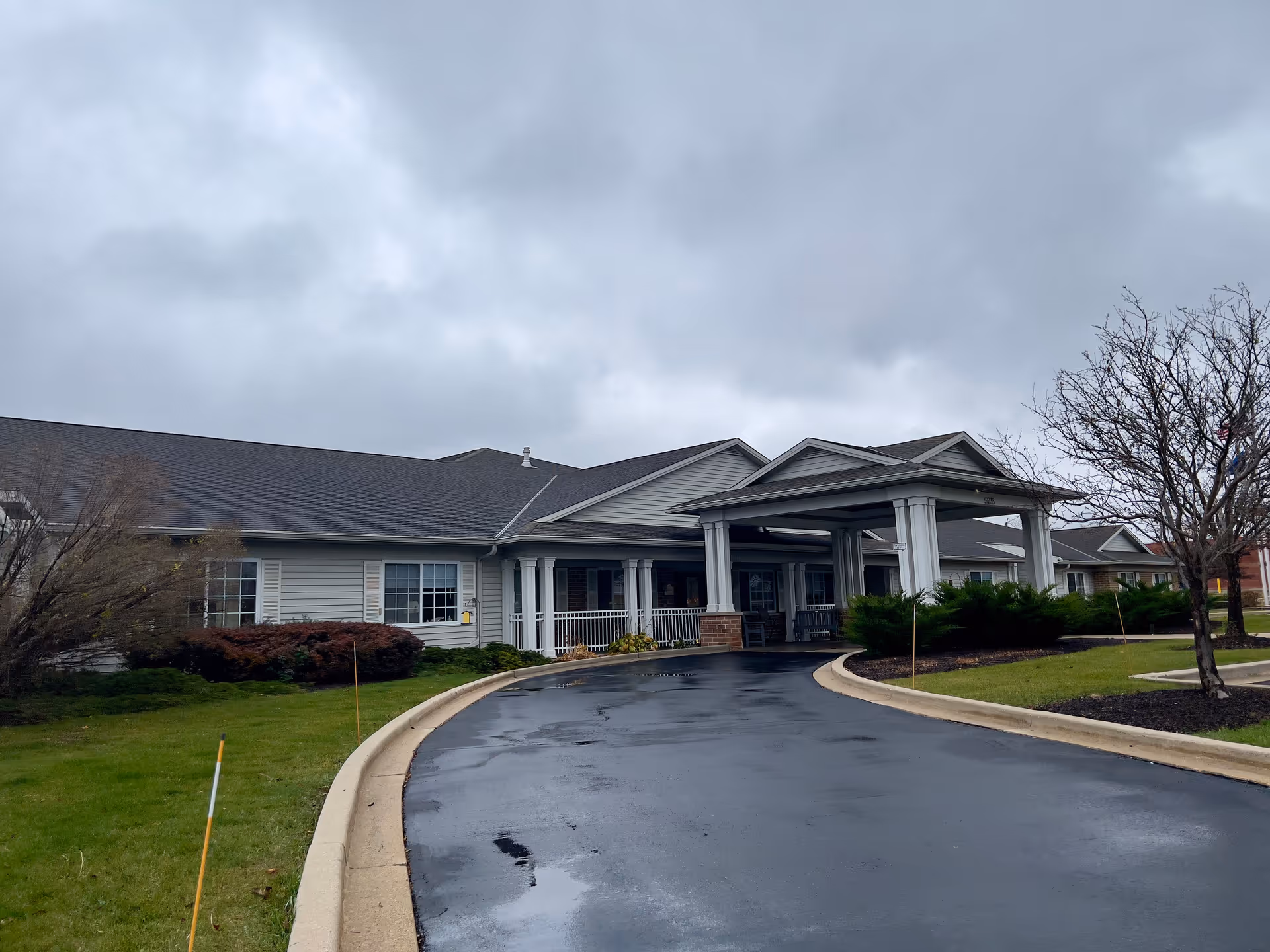 Exterior view of Brenwood Park Assisted Living facility on a cloudy day, showing a single-story building with a covered entrance, a curved driveway, green lawns, and some leafless trees.