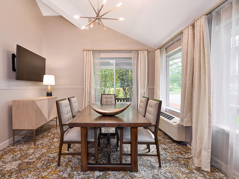 Bright dining room with a wooden table and six upholstered chairs, large windows with curtains, a sideboard and lamp, wall-mounted TV, and a modern chandelier.