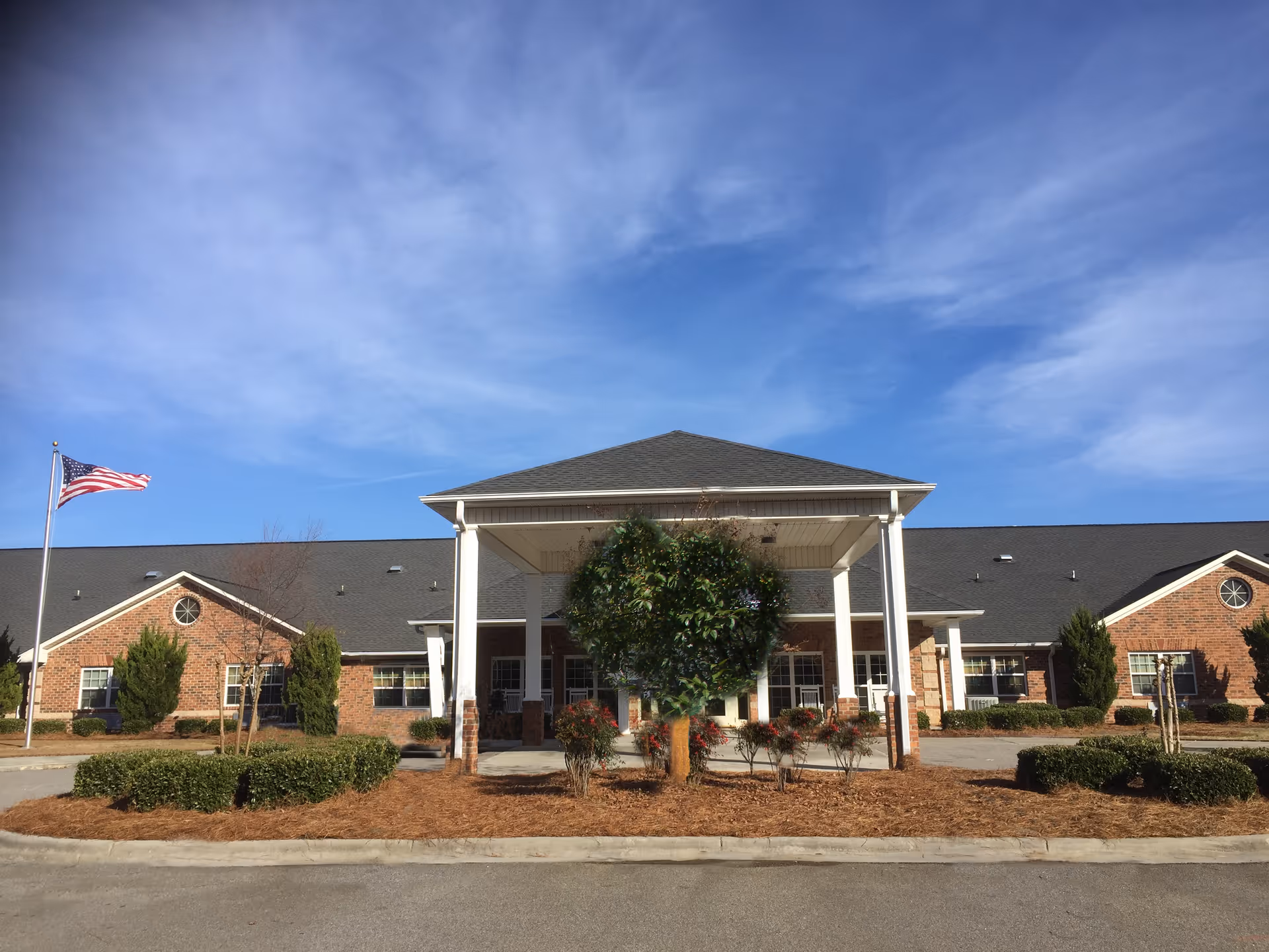 Front exterior view of a single-story brick building with a covered entrance supported by white columns. There is a landscaped area with bushes and a small tree in front of the entrance. An American flag is flying on a flagpole to the left. The sky is clear with some clouds.