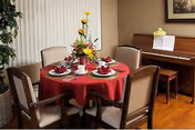 A small dining area with a round table set for four with a red tablecloth and floral centerpiece, surrounded by chairs and a piano against the wall.