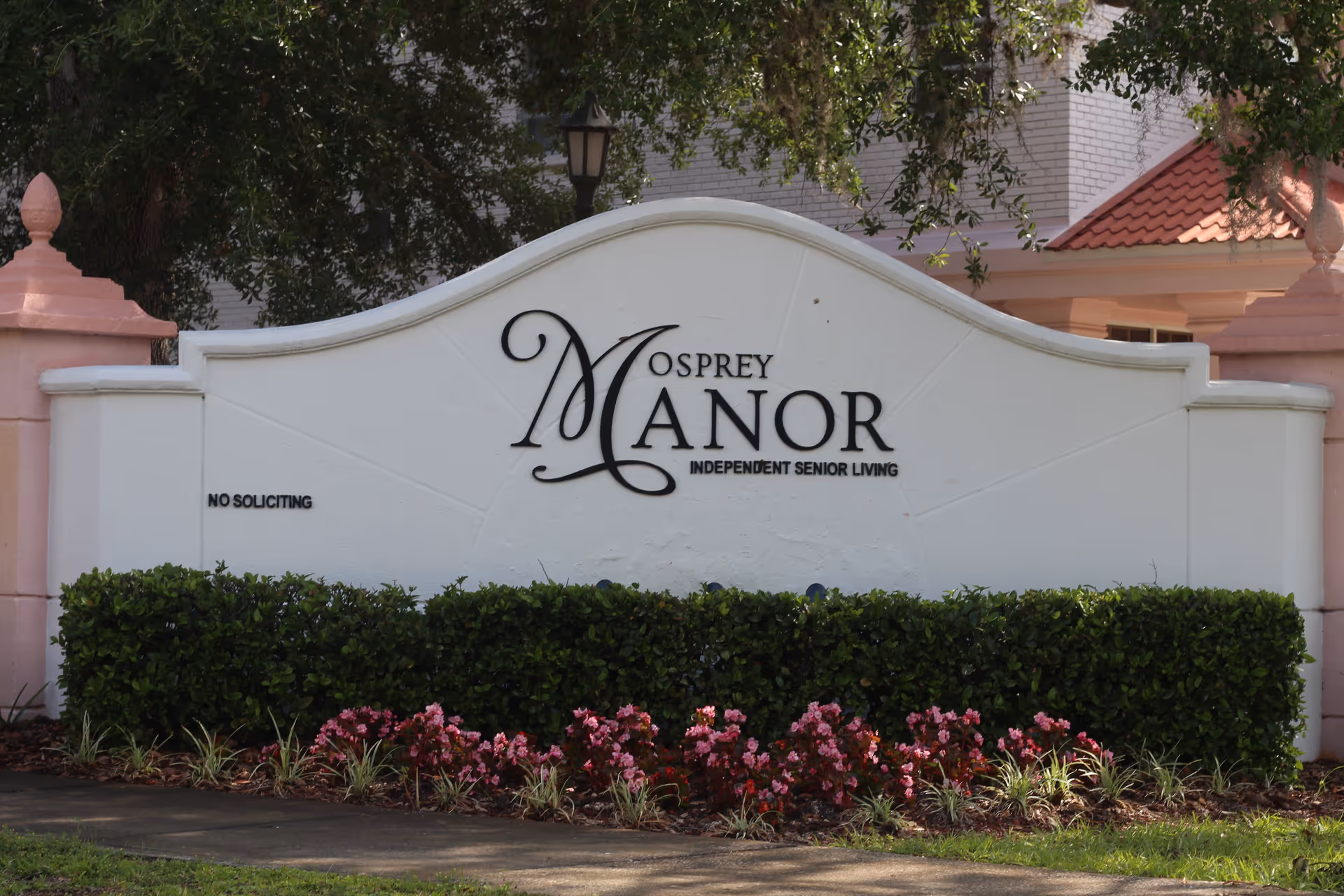 A white curved sign with black lettering that reads 'Osprey Manor Independent Senior Living' surrounded by green bushes and pink flowers, with a pink pillar and a building with a red-tiled roof in the background.