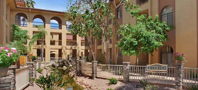 Outdoor courtyard area of a senior living facility with a small pond, stone pillars, metal railings, trees, and plants surrounded by multi-story buildings with arched windows and balconies under a clear blue sky.