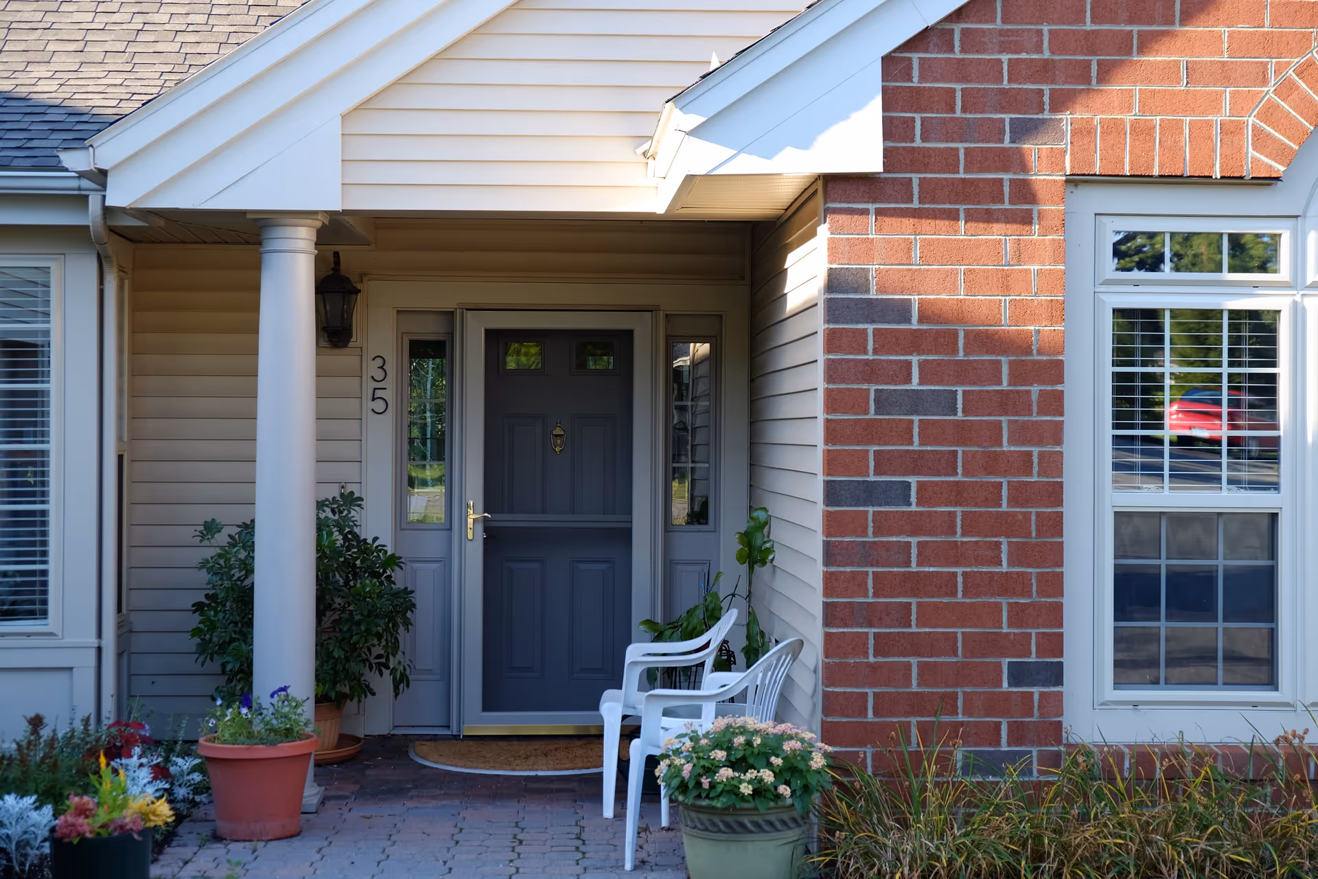 Front entrance of a residential building with a gray door numbered 35, flanked by sidelights. There are two white plastic chairs and several potted plants on the brick-paved porch. The exterior walls are a combination of beige siding and red brick, with a window on the right side.