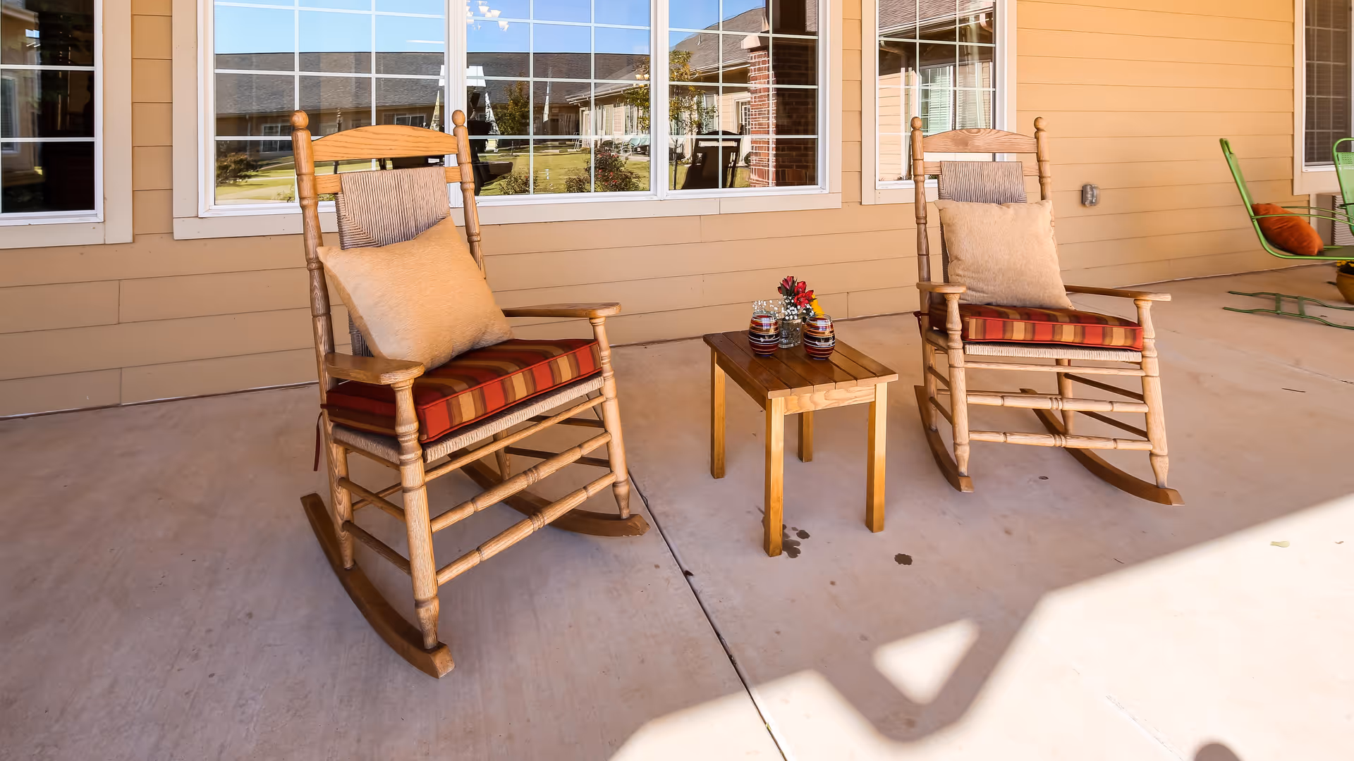 Two wooden rocking chairs with cushions and pillows are placed on a covered patio next to a small wooden table with decorative items. The patio has a beige exterior wall with large windows reflecting the outdoor area.