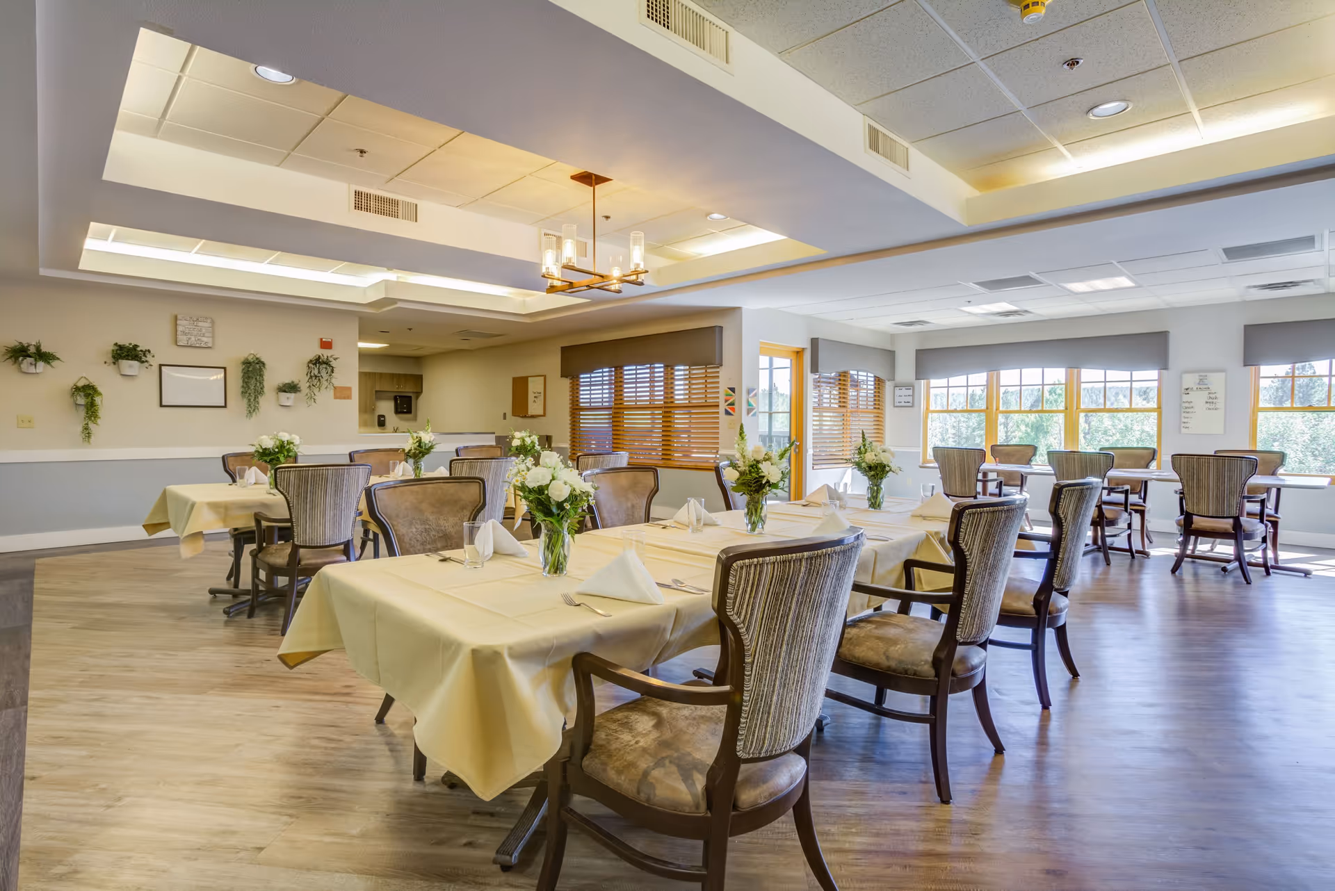 A bright and spacious dining room in a senior living community with several tables covered in light yellow tablecloths, each set with white napkins and floral centerpieces. The room features large windows with wooden blinds allowing natural light to fill the space, and comfortable upholstered chairs around the tables. The ceiling has recessed lighting and a decorative chandelier.