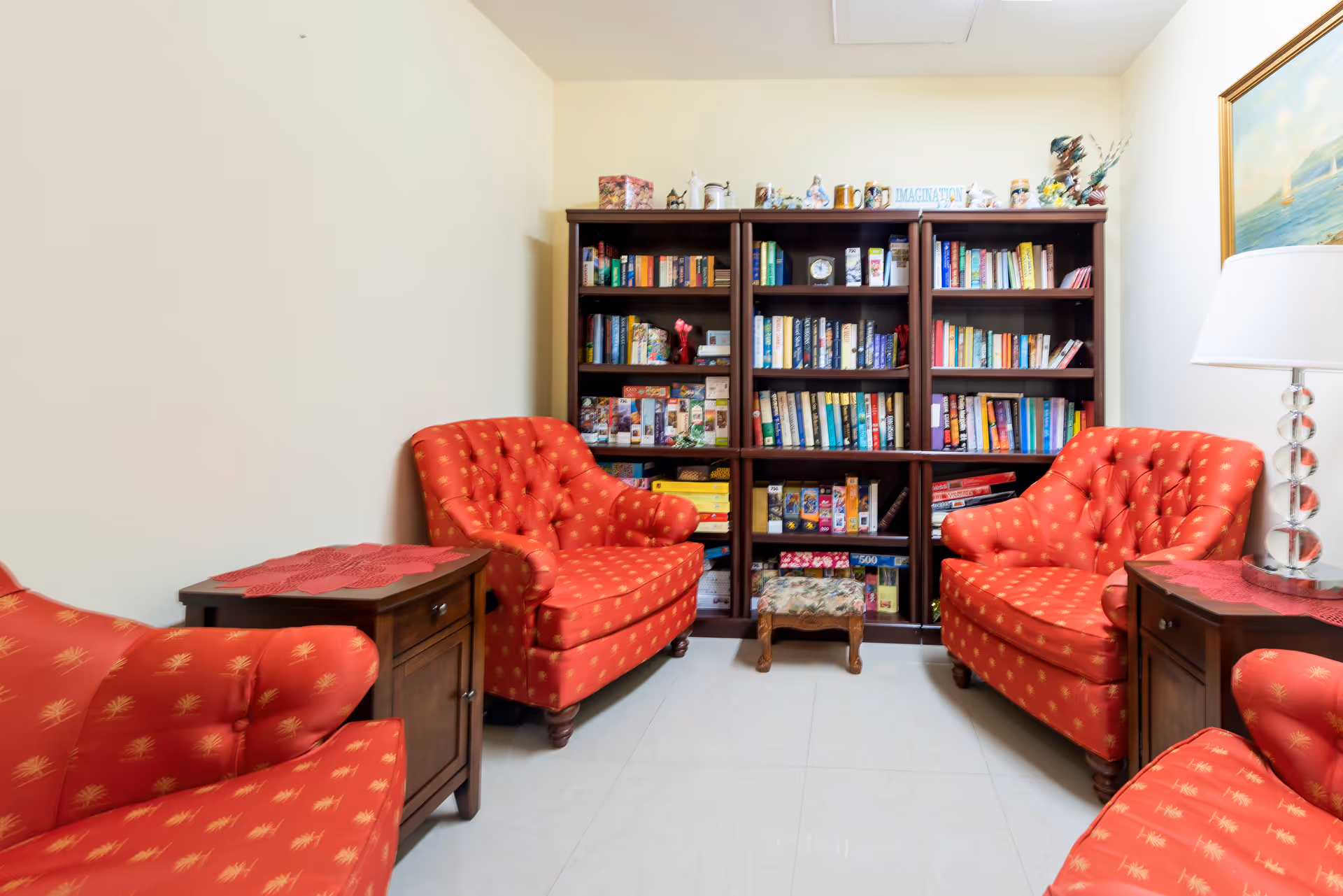 A cozy sitting area with four red upholstered armchairs arranged around two wooden side tables with red doilies. Behind the chairs is a large dark wooden bookshelf filled with books, board games, and decorative items. A floor lamp with a white shade stands to the right, and a framed painting hangs on the wall above it.