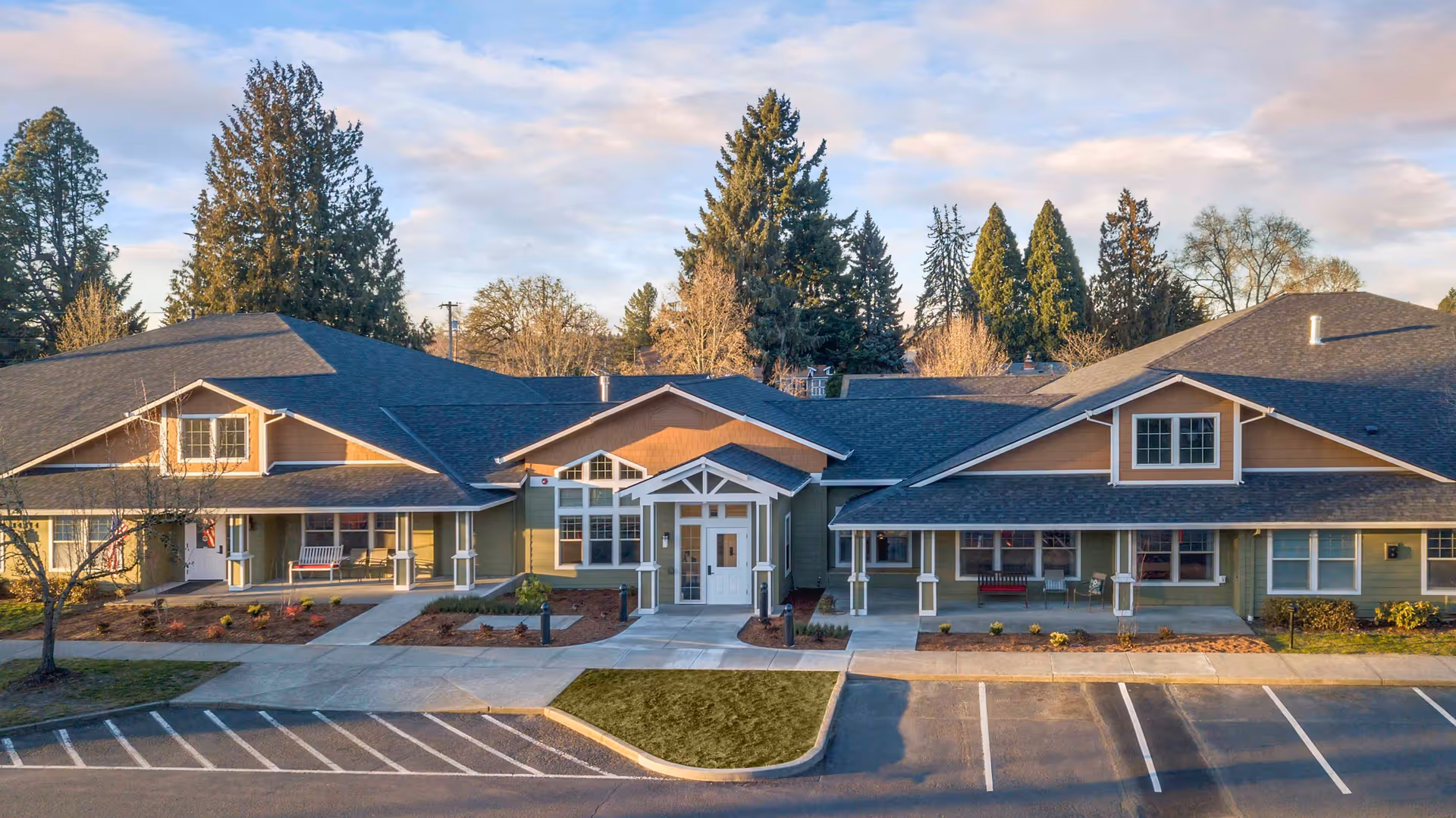 Exterior view of a single-story senior living facility building with a peaked roof, multiple windows, and a central entrance with a small covered porch. The building is surrounded by a parking lot and landscaped areas with small plants and trees, under a partly cloudy sky.