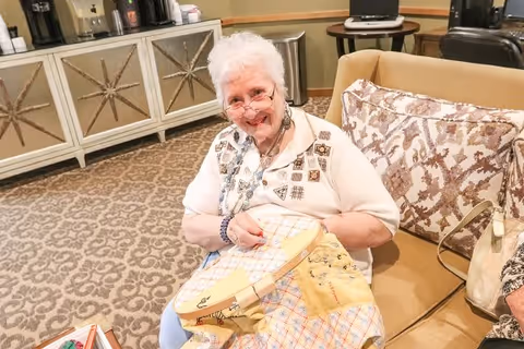 An elderly woman with white hair and glasses is sitting on a beige couch in a cozy room. She is smiling and engaged in embroidery work on a large piece of fabric. Behind her is a cabinet with a star design and some coffee machines on top. The room has patterned carpet and warm lighting.