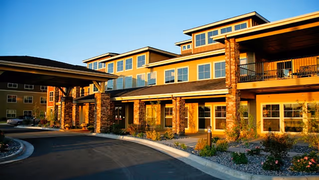 Exterior view of a multi-story senior living facility building with large windows, stone pillars, and a covered entrance, surrounded by landscaped plants and a curved driveway under a clear blue sky.