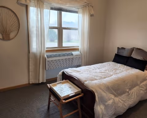 A simple senior bedroom with a single bed dressed in a white comforter, a small tray table at the foot, and a curtained window above a wall air-conditioning unit.