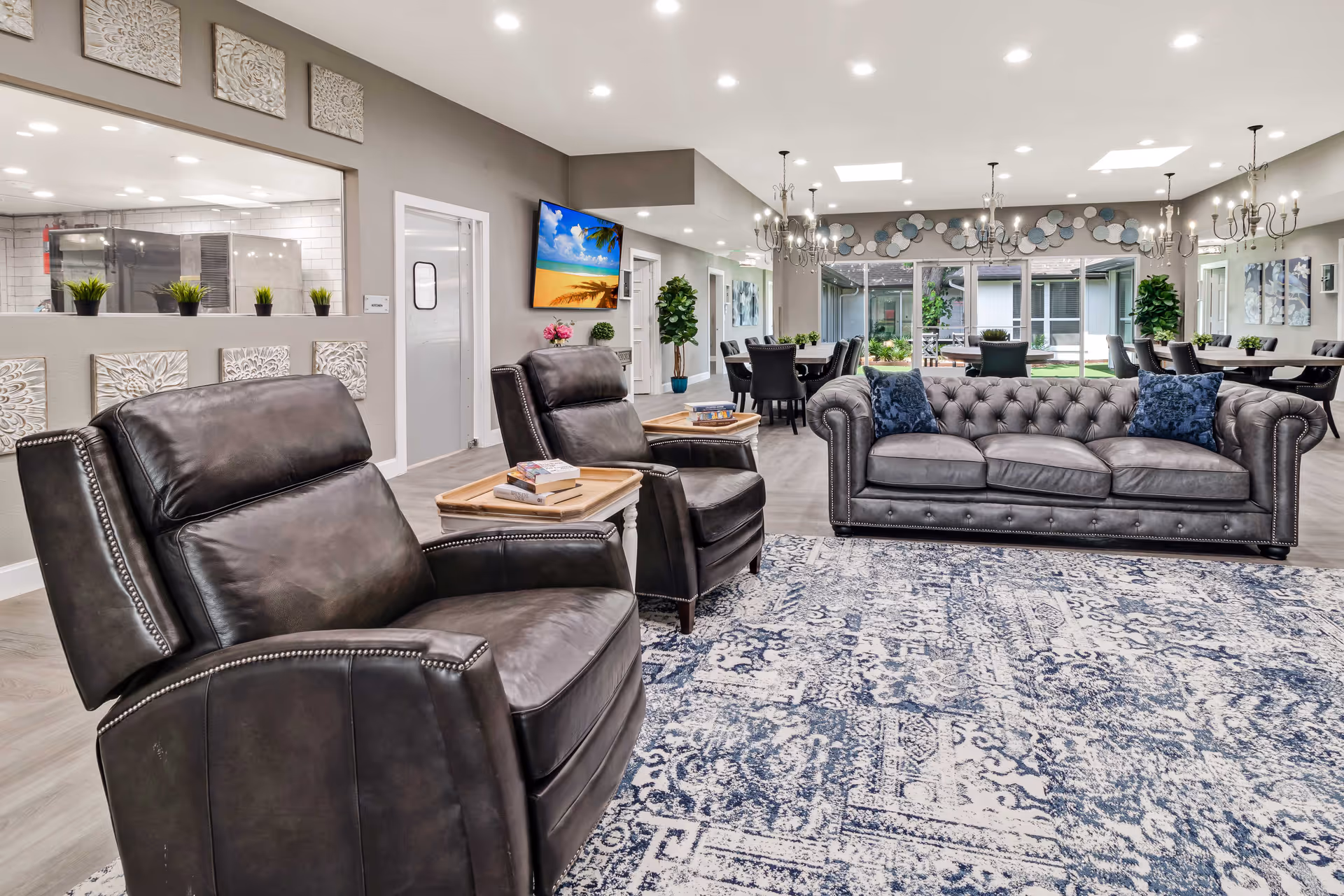 A spacious and well-lit living room area in an assisted living facility featuring two dark leather recliners and a matching tufted leather sofa with blue pillows. The room has a large patterned rug, several dining tables with chairs in the background, modern chandeliers, and large windows letting in natural light. Decorative wall art and plants add to the inviting atmosphere.