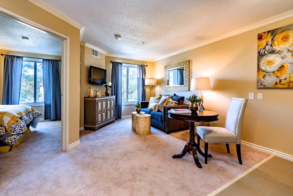 A cozy senior living interior featuring a small living room with a dark blue couch adorned with patterned pillows, a round wooden table with a white cushioned chair, a decorative mirror on the wall, and a cabinet with a mounted TV. To the left, an open doorway reveals a bedroom with a bed covered in a yellow and gray patterned comforter. Both rooms have large windows with blue curtains letting in natural light.