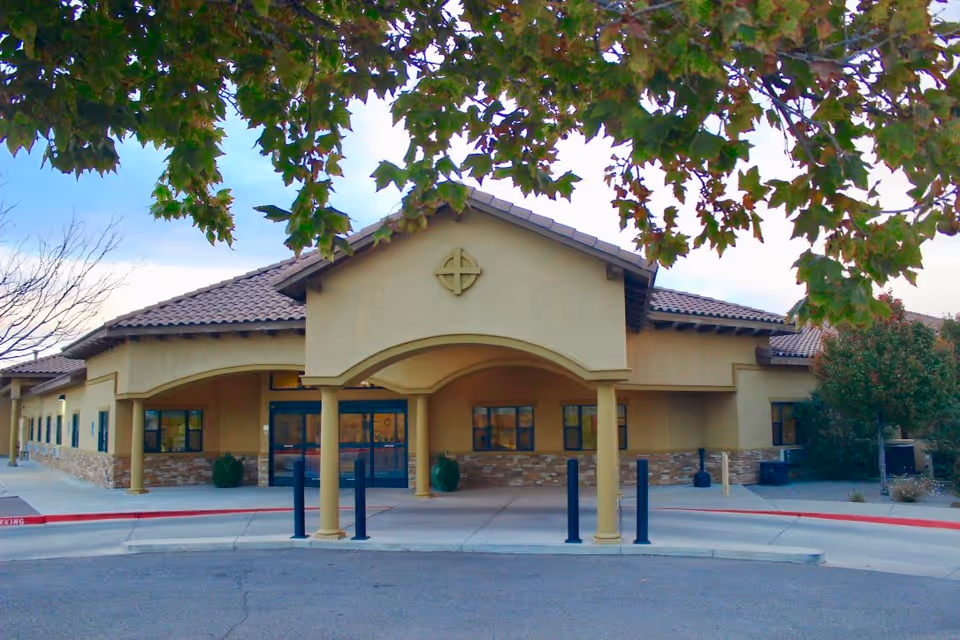 Front exterior view of a single-story assisted living facility building with a covered entrance supported by columns, beige walls, stone accents, and a tiled roof. Trees with green leaves frame the top of the image.