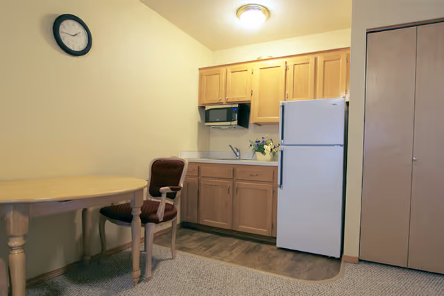 Compact kitchenette in a senior apartment with wooden cabinets, a white refrigerator, a round table and an upholstered chair.