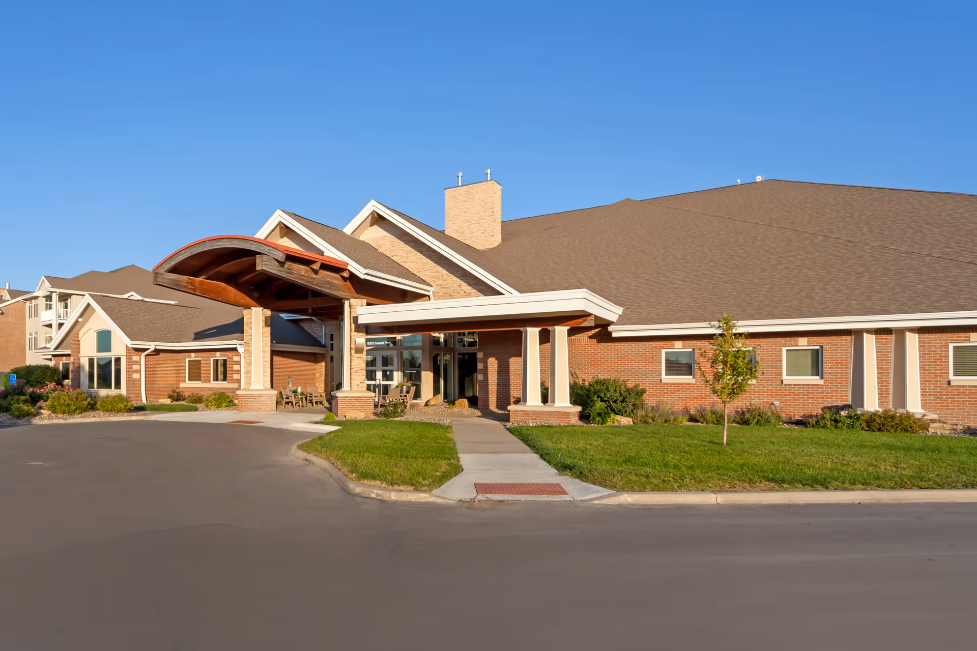 Exterior view of Elk Ridge Village Assisted Living building with a covered entrance, brick walls, large windows, and a well-maintained lawn under a clear blue sky.