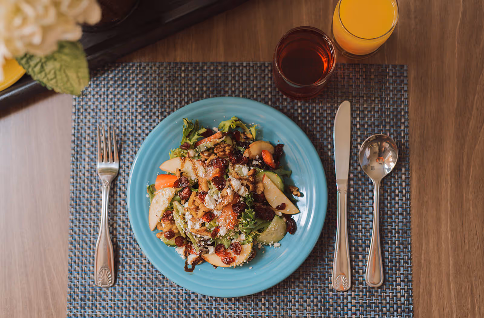 A blue plate with a fresh salad containing mixed greens, sliced apples, walnuts, dried cranberries, and crumbled cheese on a woven placemat. There is a fork on the left side of the plate, and a knife and spoon on the right side. Two glasses with orange juice and a dark red beverage are placed above the plate on a wooden table.