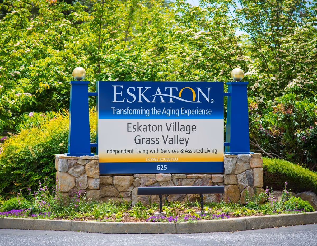 A large blue and white sign for Eskaton Village Grass Valley, an independent living and assisted living facility, set in a landscaped area with green bushes, flowering plants, and trees in the background.