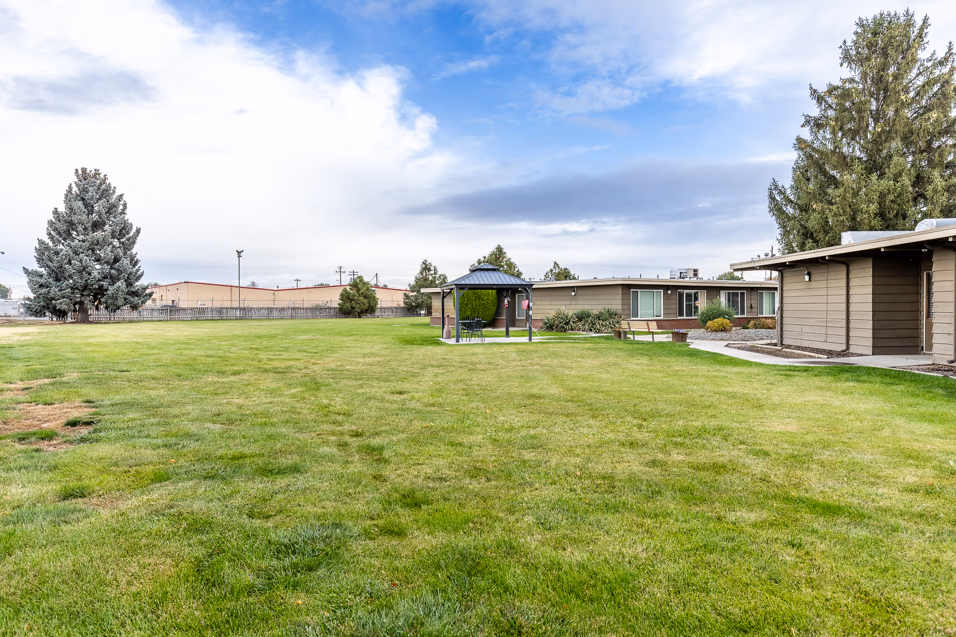Wide grassy courtyard with a gazebo and single-story buildings under a partly cloudy sky.