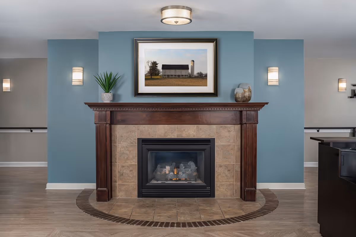 A cozy indoor fireplace with a wooden mantel and beige tiles surrounding the firebox. Above the mantel is a framed picture of a barn and silo in a rural setting. The wall behind the fireplace is painted blue, and there are two wall sconces on either side. A small potted plant and decorative vases are placed on the mantel.