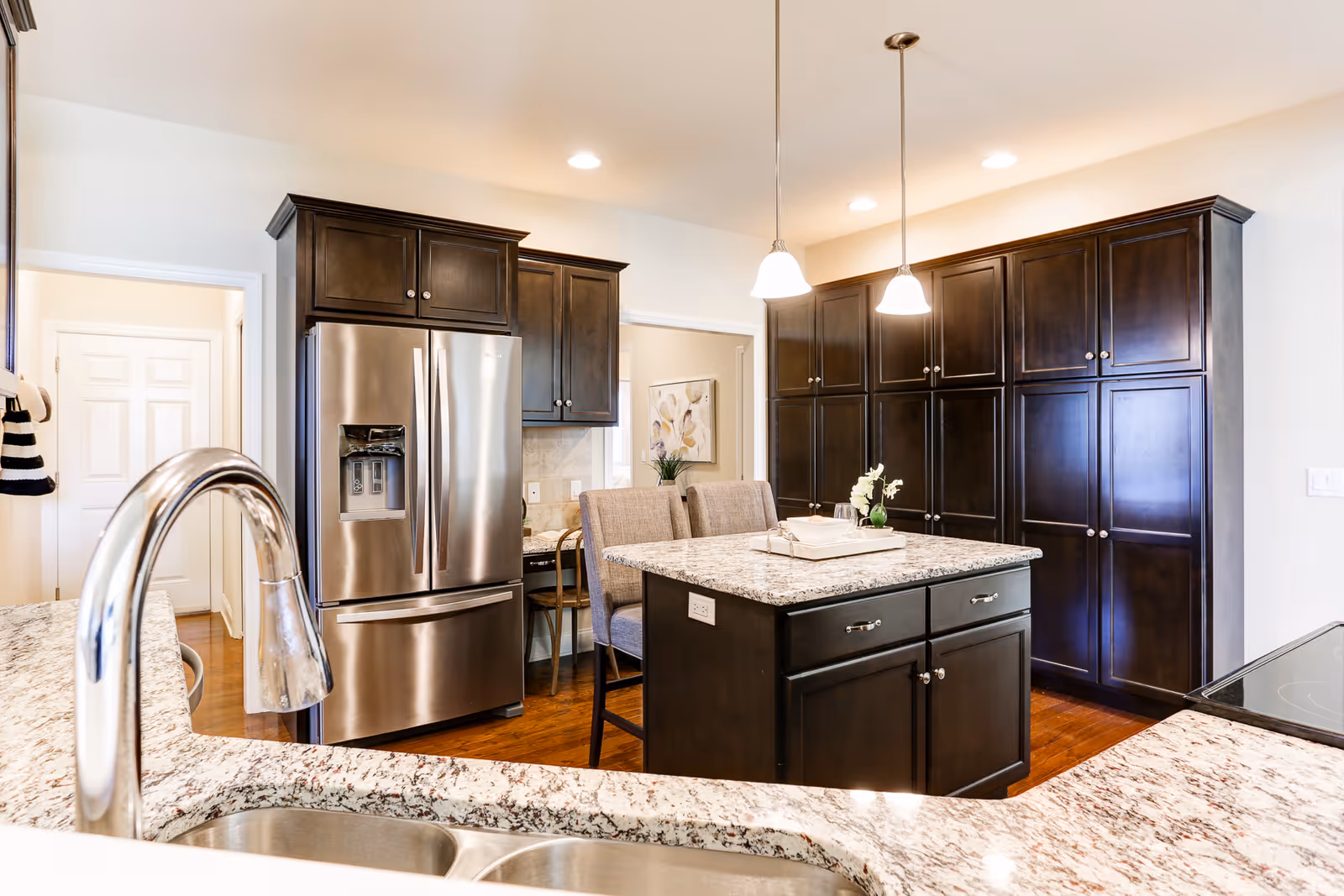 Bright modern kitchen with granite countertops, a center island, stainless steel refrigerator, dark wood cabinets, pendant lights, and a sink in the foreground.