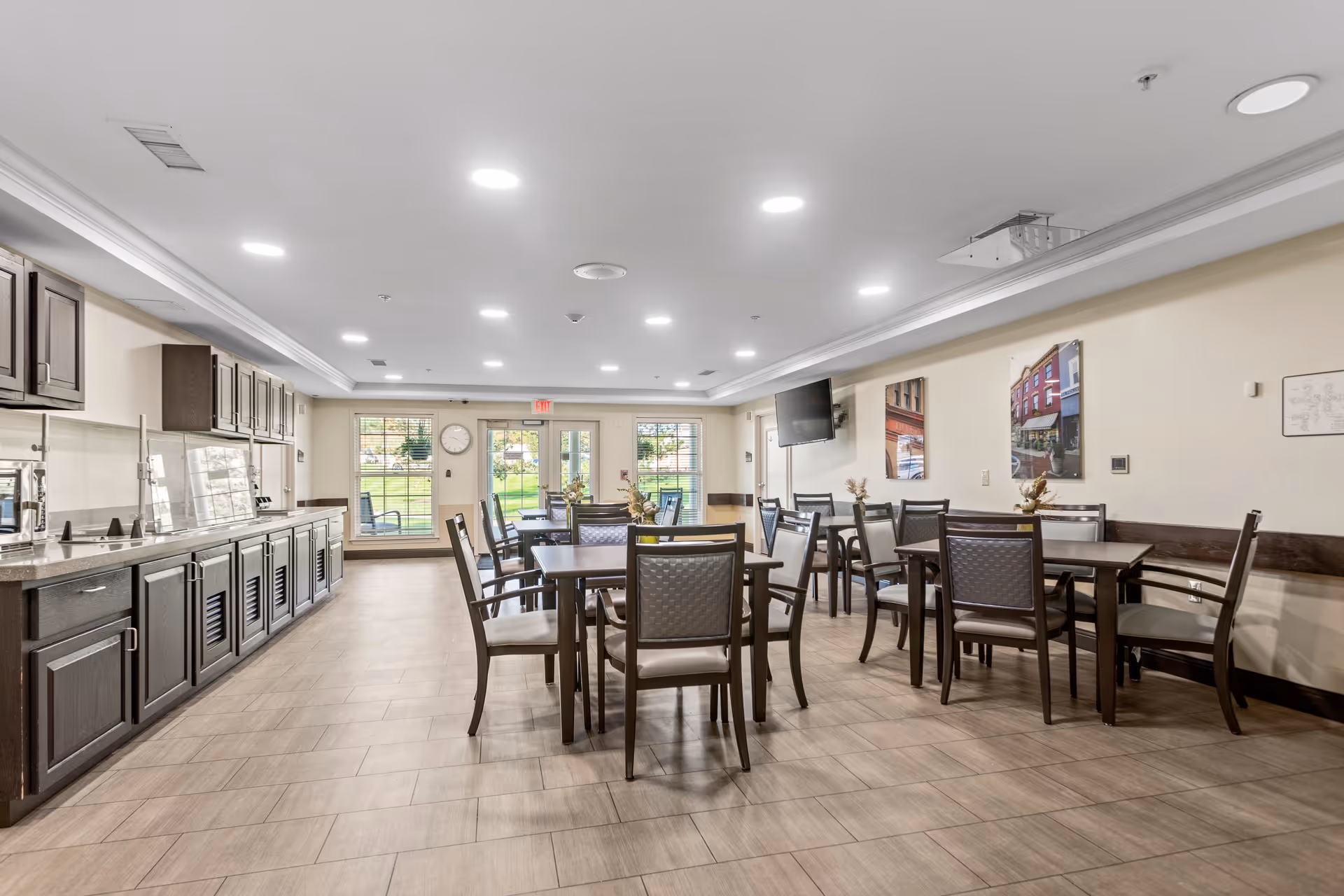 A bright dining room with several dark wood tables and chairs arranged neatly. The room features a long counter with cabinets on the left side, a wall-mounted TV on the right, and large windows and glass doors at the far end letting in natural light. The floor is tiled, and the ceiling has recessed lighting.
