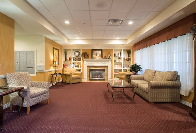 A cozy senior living facility common area with a red carpet, beige patterned sofa, two beige armchairs, a glass coffee table, and a fireplace with built-in shelves decorated with books and ornaments. Large windows with sheer white curtains and a brown valance allow natural light into the room.