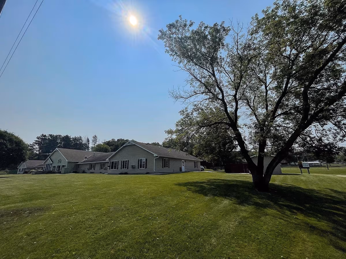 A sunny outdoor view of a single-story building with beige siding and multiple windows, surrounded by a large green lawn and a tall tree casting a shadow. The sky is clear and blue with the sun shining brightly. A sign in the background reads 'Park Lane Estates.'