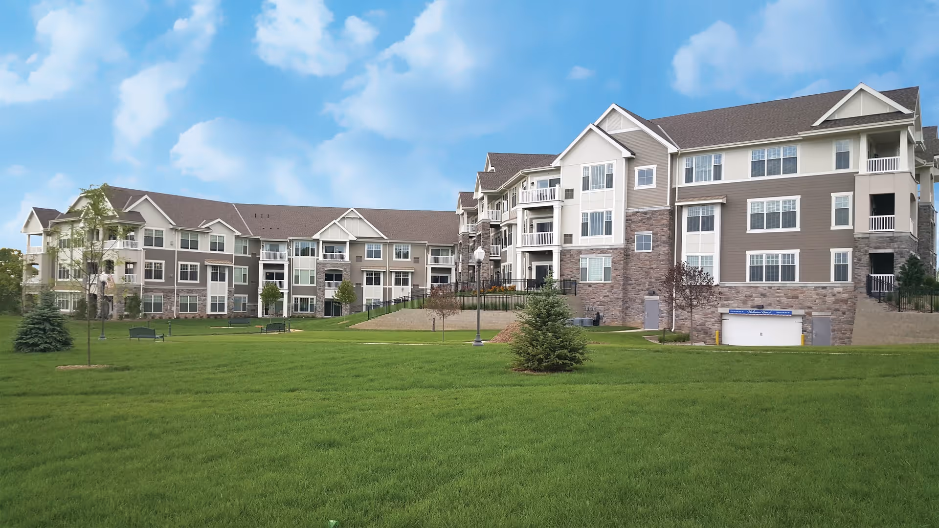 Exterior view of a large, multi-story senior living facility building with beige and stone facade, surrounded by a well-maintained green lawn and small trees under a partly cloudy blue sky.