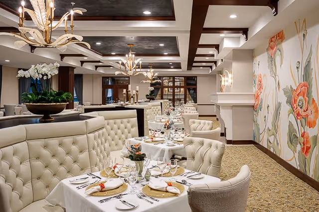 Elegant dining room with tufted cream-colored chairs and booths, tables set with white tablecloths, plates, glasses, and floral centerpieces. The room features decorative ceiling beams, chandeliers, and a large floral mural on one wall.