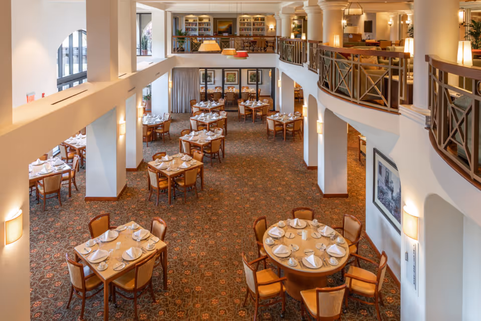 Spacious dining room with multiple tables set with white napkins, plates, and cups. The room features a patterned carpet, warm lighting, and a second-floor balcony with seating and bookshelves. Large windows allow natural light to fill the space.