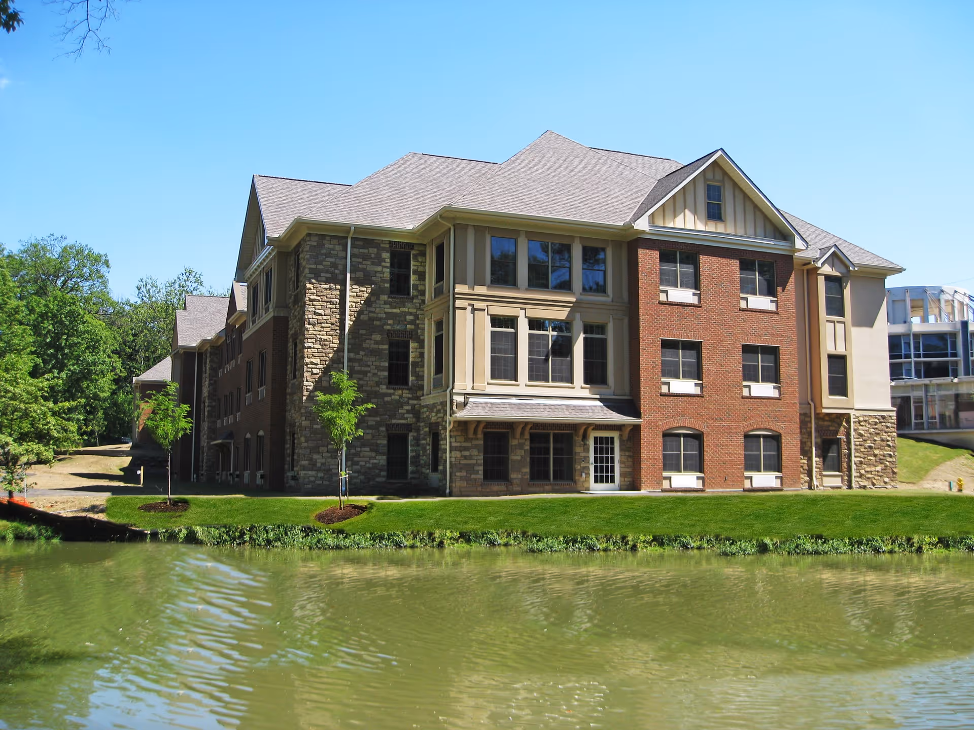 A multi-story senior living facility building with a combination of brick and stone exterior walls, large windows, and a pitched roof, situated next to a pond with green grass and small trees around it under a clear blue sky.