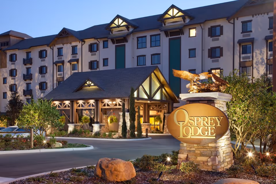 Exterior view of a large multi-story senior living facility building at dusk with warm lights illuminating the entrance area. In the foreground, there is a stone sign with a sculpture of two birds and the text 'Osprey Lodge'. The building has a pitched roof and multiple windows with decorative shutters.