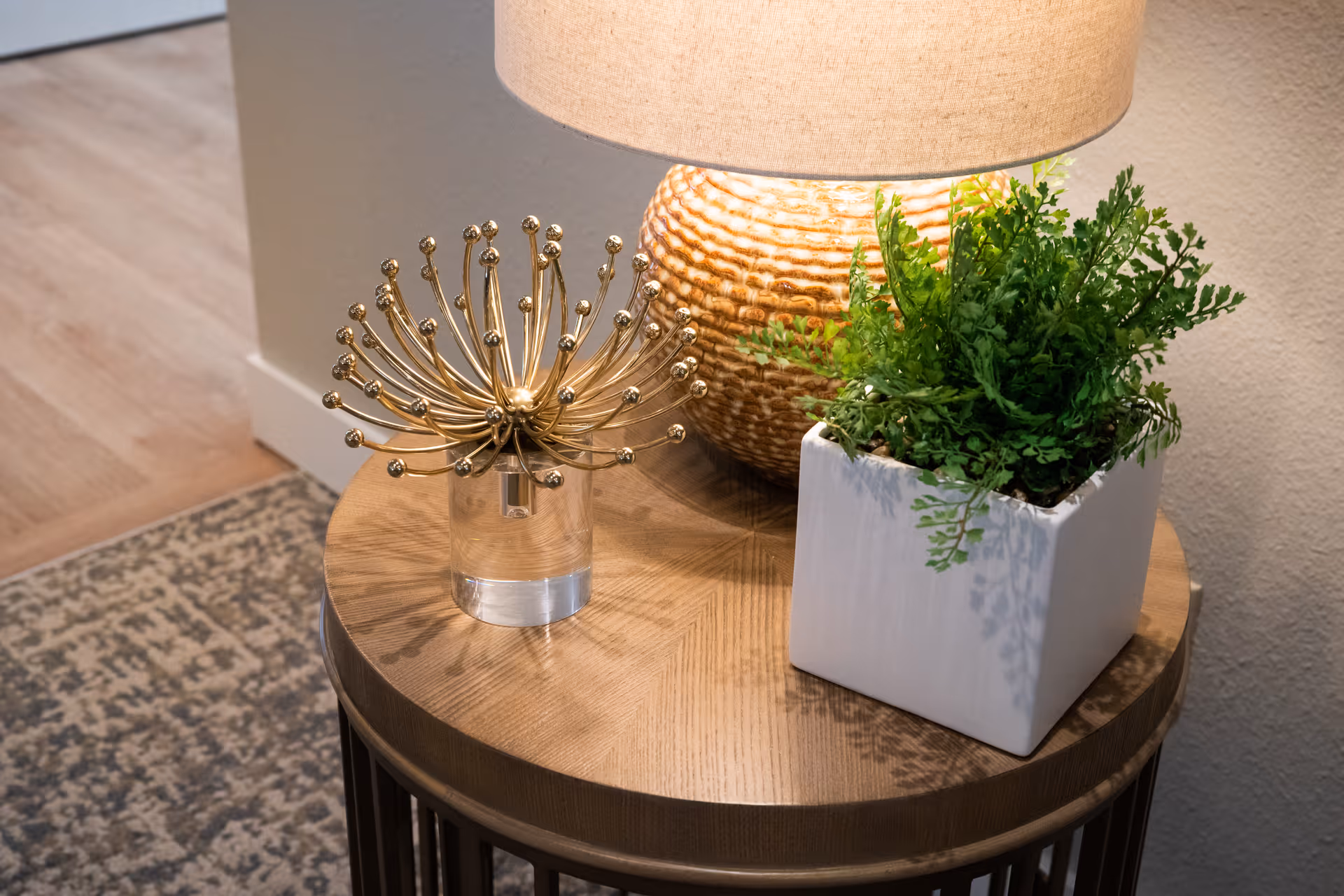 Round side table with a lamp, a gold decorative sculpture, and a potted fern next to a rug and hardwood floor.