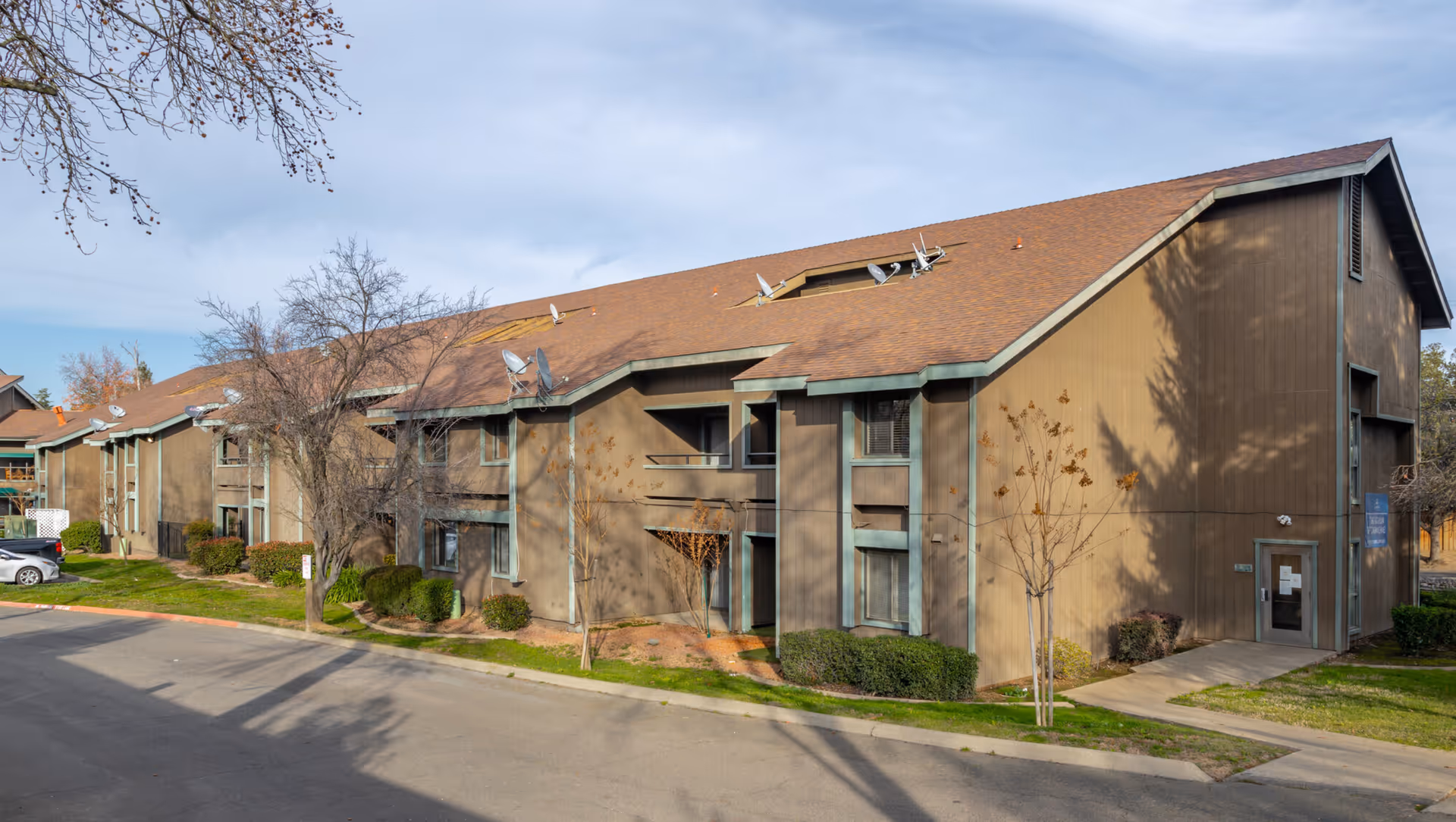 Exterior view of a two-story brown residential building with a sloped roof and multiple satellite dishes mounted on top. The building is surrounded by leafless trees, bushes, and a paved driveway with parked cars. The sky is partly cloudy.