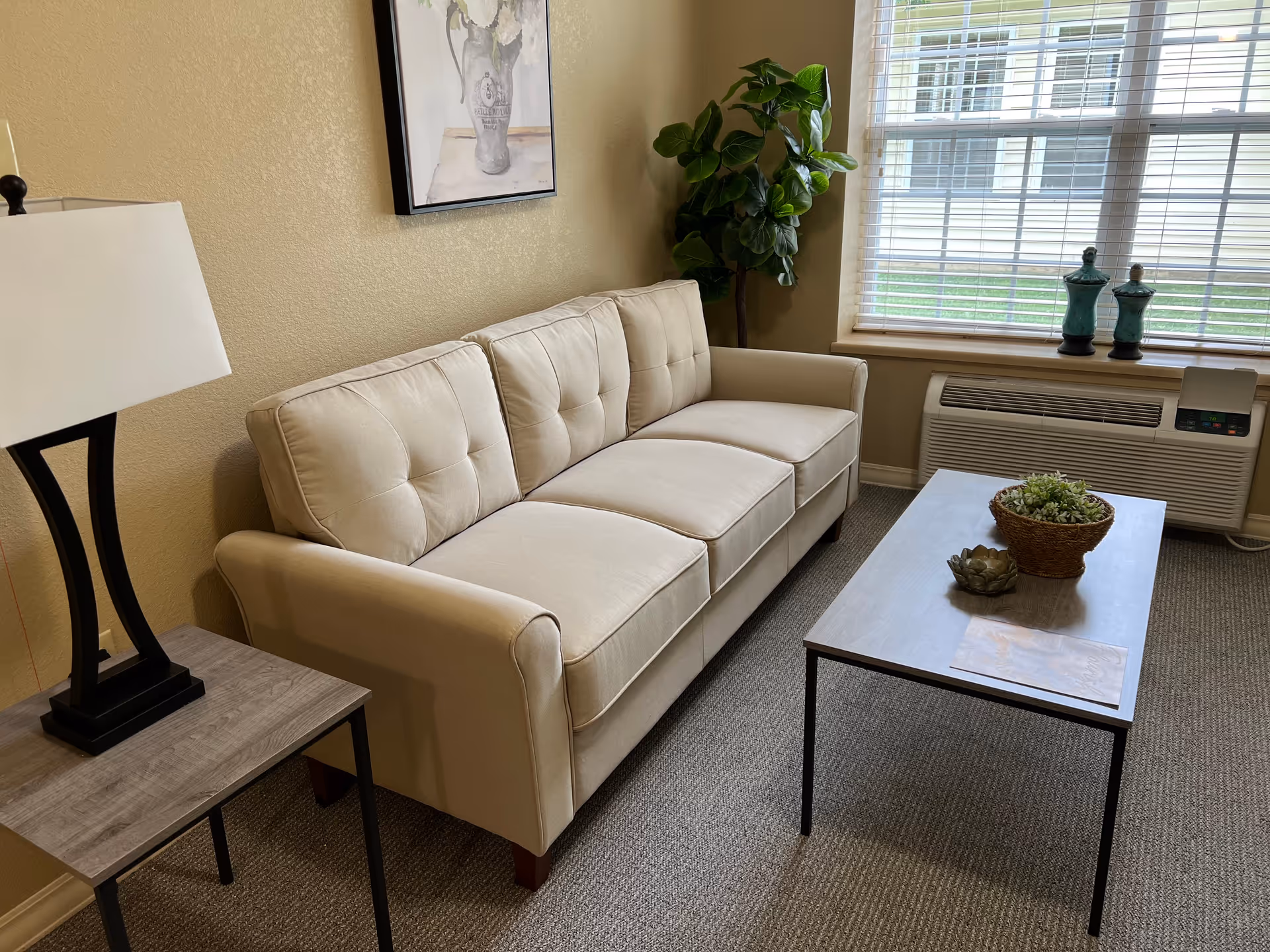 A cozy living room area with a beige three-seater sofa, a wooden side table with a lamp, a rectangular coffee table with decorative plants, a large window with blinds, and a green potted plant in the corner.