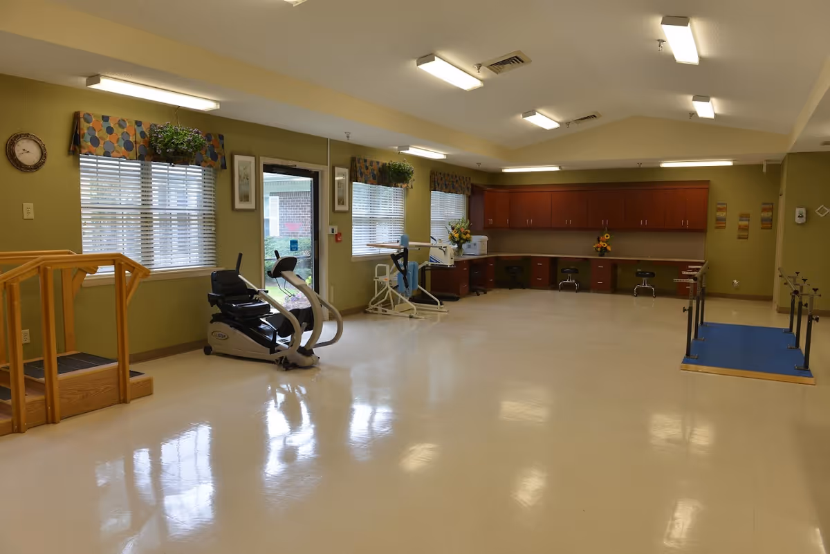 A spacious rehabilitation or physical therapy room with exercise equipment including a stationary bike and parallel bars. The room has green walls, large windows with patterned valances, wooden cabinets along the back wall, and a small set of stairs with handrails on the left side.