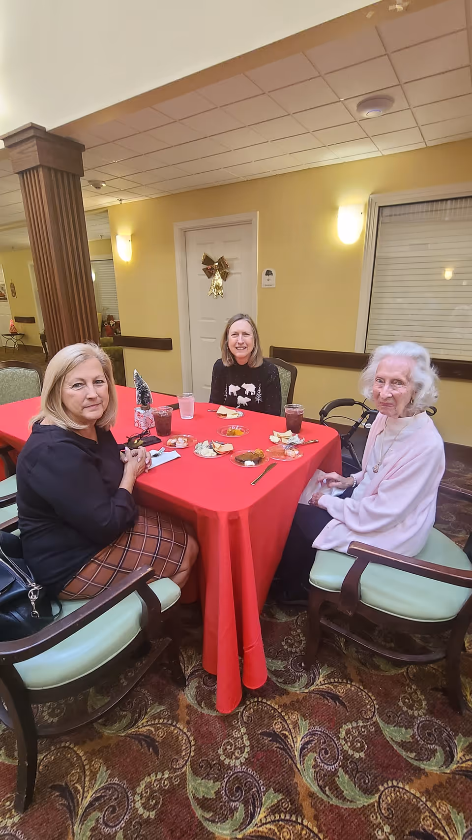 Three women sitting around a red tablecloth-covered table in a senior living community dining area. They have plates with food and drinks in front of them. The room has yellow walls, a decorative column, and a door with a holiday decoration.
