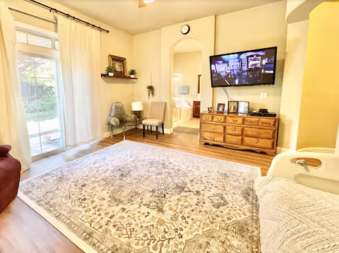A bright living room with a large patterned area rug, a wooden dresser with a flat-screen TV mounted above it, two chairs, a glass side table with a lamp, a wall shelf with plants and a picture frame, and a large window with white curtains letting in natural light. The room has light-colored walls and wooden flooring.