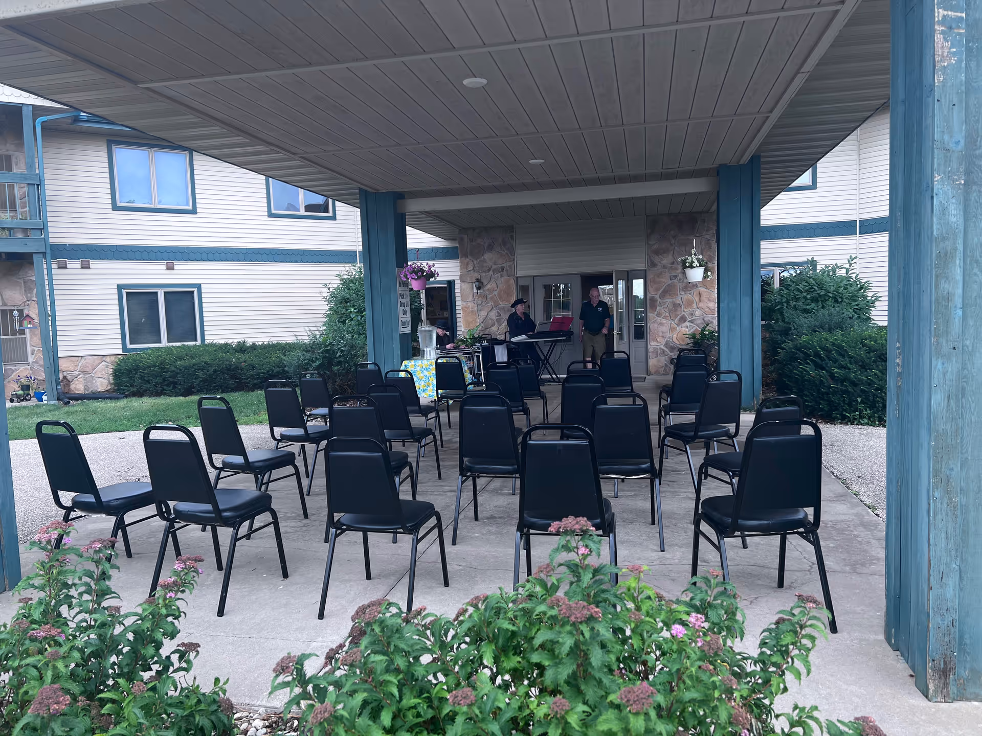 Outdoor covered seating area with rows of black chairs arranged facing a small table with a keyboard and two people standing behind it. The area is part of a building with stone and siding exterior walls, surrounded by greenery and flowering plants.