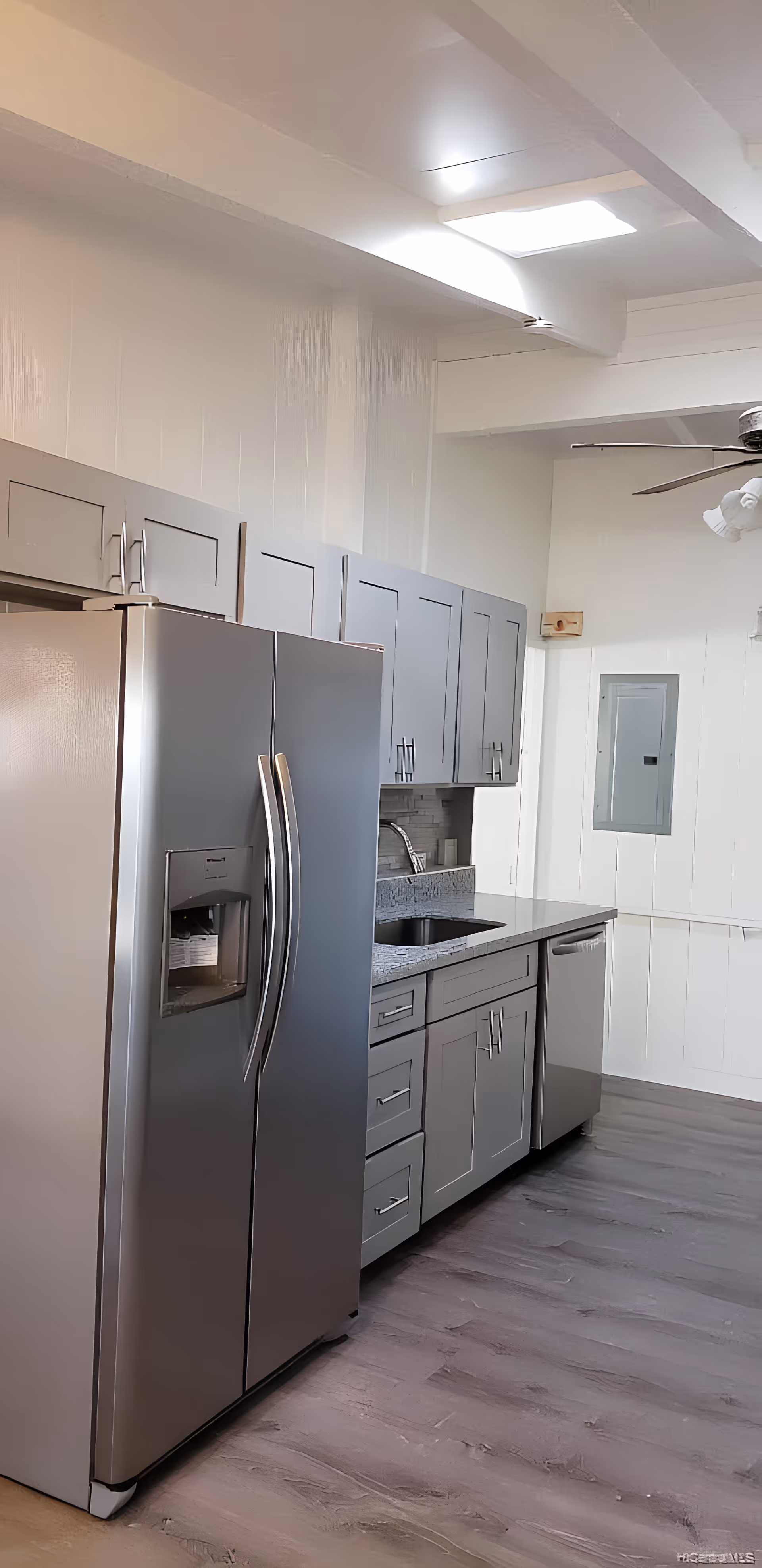 Modern kitchen with stainless steel refrigerator, dishwasher, and sink. Grey cabinets with silver handles line the wall above and below the countertop. The floor is a light wood laminate, and a ceiling fan with lights is visible. There is a skylight providing natural light.