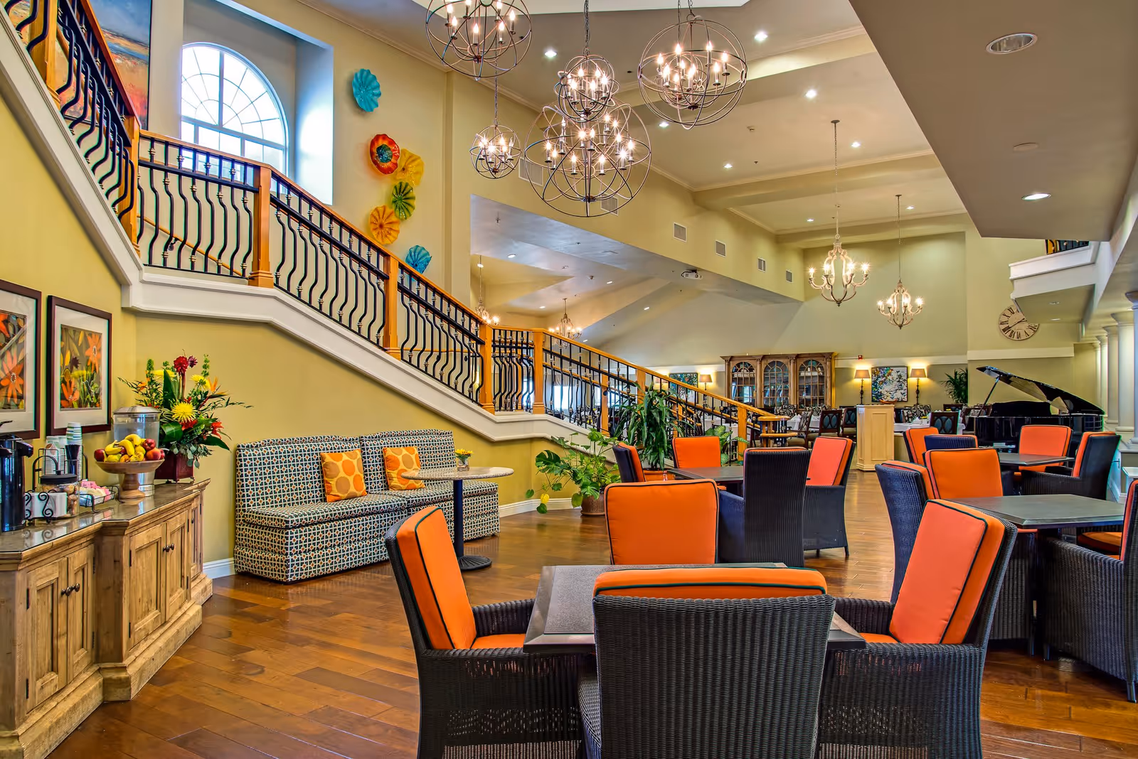 A spacious and well-lit common area with multiple tables and chairs featuring orange cushions. The room has wooden flooring, a staircase with a decorative railing, colorful wall decorations, and several chandeliers hanging from the ceiling. There is a sideboard with fruit and flowers, and a grand piano is visible in the background.
