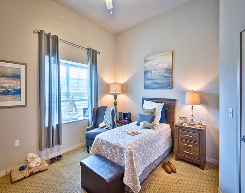 A cozy bedroom with a single bed covered in a patterned beige and white bedspread, a wooden headboard, and several pillows including a decorative one. Next to the bed is a wooden nightstand with a lamp, a clock, and some small items. A blue upholstered armchair with a hat and a blanket is placed near a window with gray curtains. On the floor near the window is a dog bed with a stuffed animal and a couple of dog bowls. The walls are light-colored with two pieces of artwork hanging, one above the bed and one beside the window. The room has a beige carpet and a ceiling fan above.