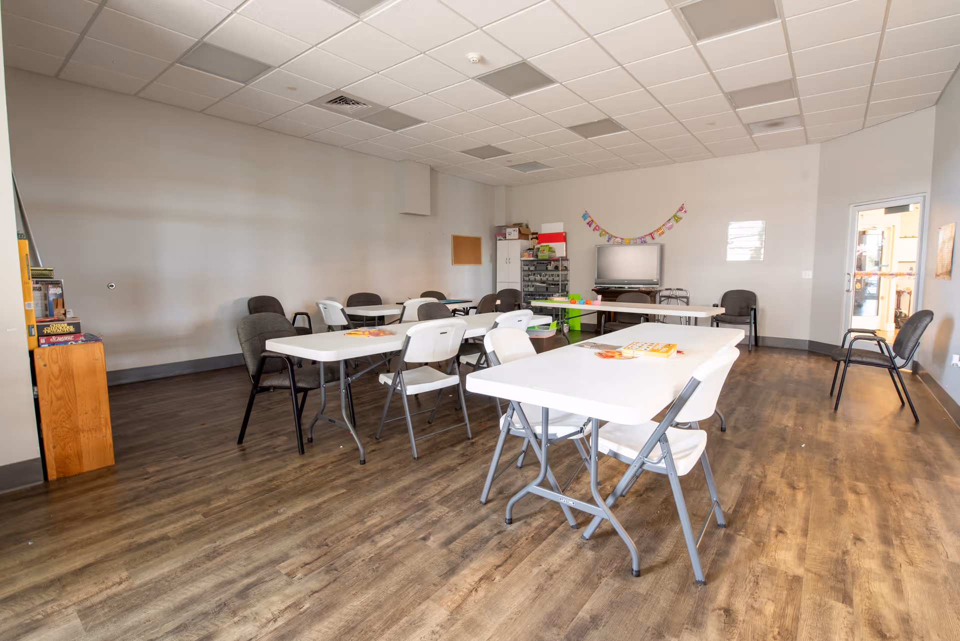 A spacious room with several white folding tables and chairs arranged in a U-shape. The room has wood flooring, a drop ceiling with fluorescent lights, and neutral-colored walls. There is a TV on a stand against the far wall with a colorful 'Happy Birthday' banner hanging above it. Various board games and supplies are visible on shelves and a small cabinet in the corner. The room appears to be a communal activity or meeting space.