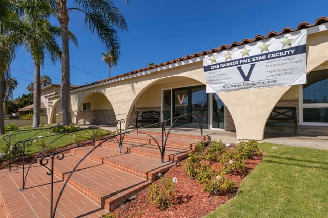 Exterior view of Victoria Healthcare and Rehabilitation facility showing a beige stucco building with arched entryways, a tiled roof, brick steps with black metal railings, palm trees, and a banner highlighting the facility's five-star CMS ranking.