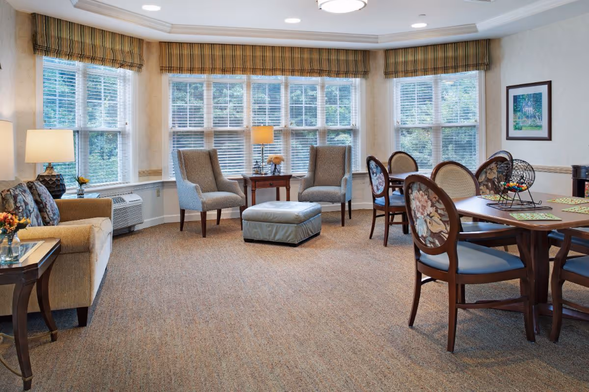 A bright and cozy common area in a senior living facility with large windows covered by blinds and plaid valances. The room features a beige sofa with floral cushions, two gray armchairs with a small wooden table and lamp between them, a padded ottoman, and a wooden dining table with floral upholstered chairs. The carpet is a neutral tone, and the walls are light-colored with a framed picture hanging.