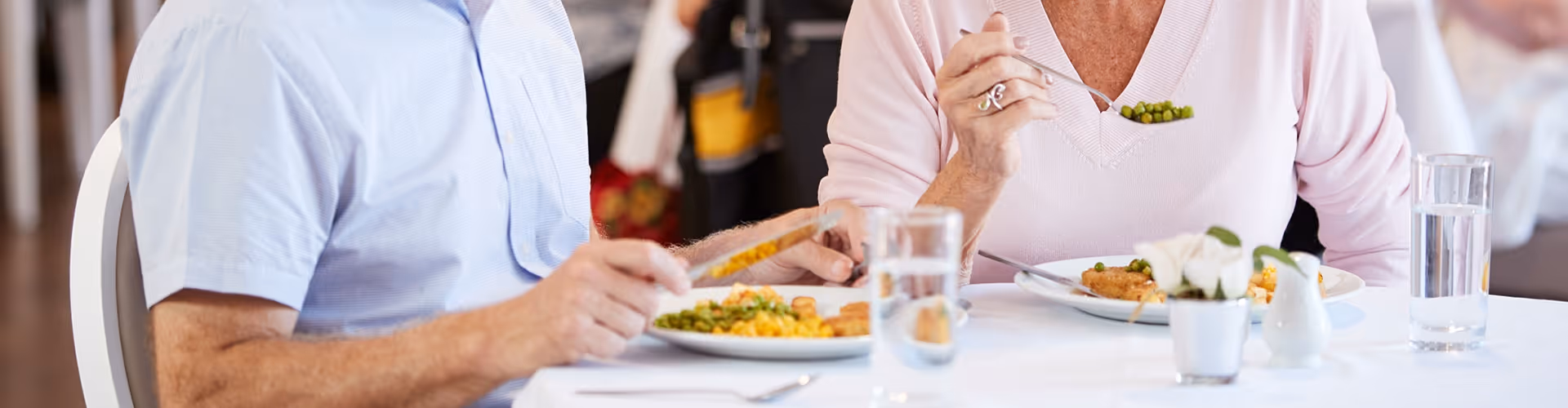 Two people seated at a dining table eating plated meals with glasses of water and a small floral centerpiece.